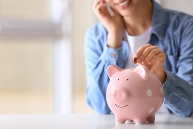 Photo of Woman putting coin into piggy bank at table indoors, closeup. Space for text