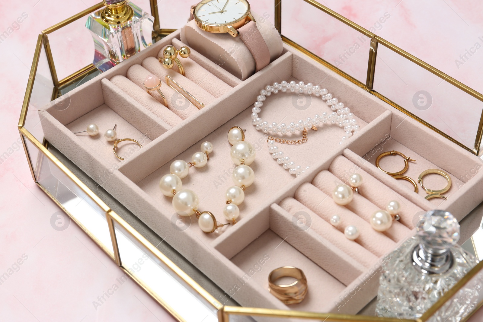 Photo of Jewelry box with many different accessories and perfumes on pink marble table, closeup