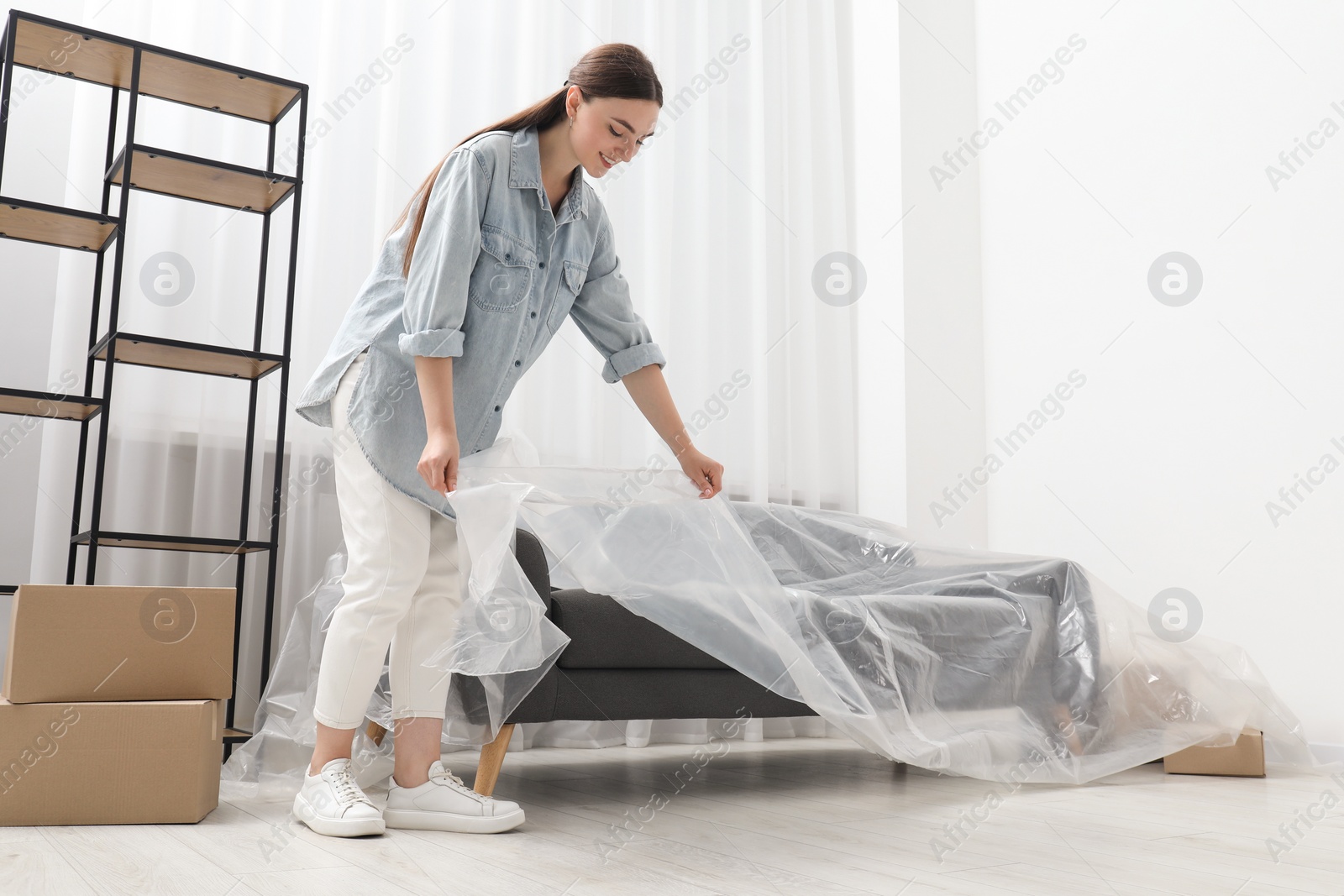 Photo of Young woman covering sofa with plastic film at home, low angle view