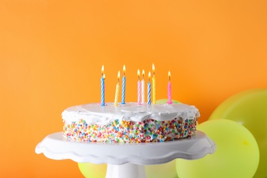 Tasty birthday cake with burning candles and balloons on orange background