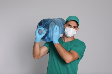 Photo of Courier in medical mask holding bottle for water cooler on light grey background. Delivery during coronavirus quarantine