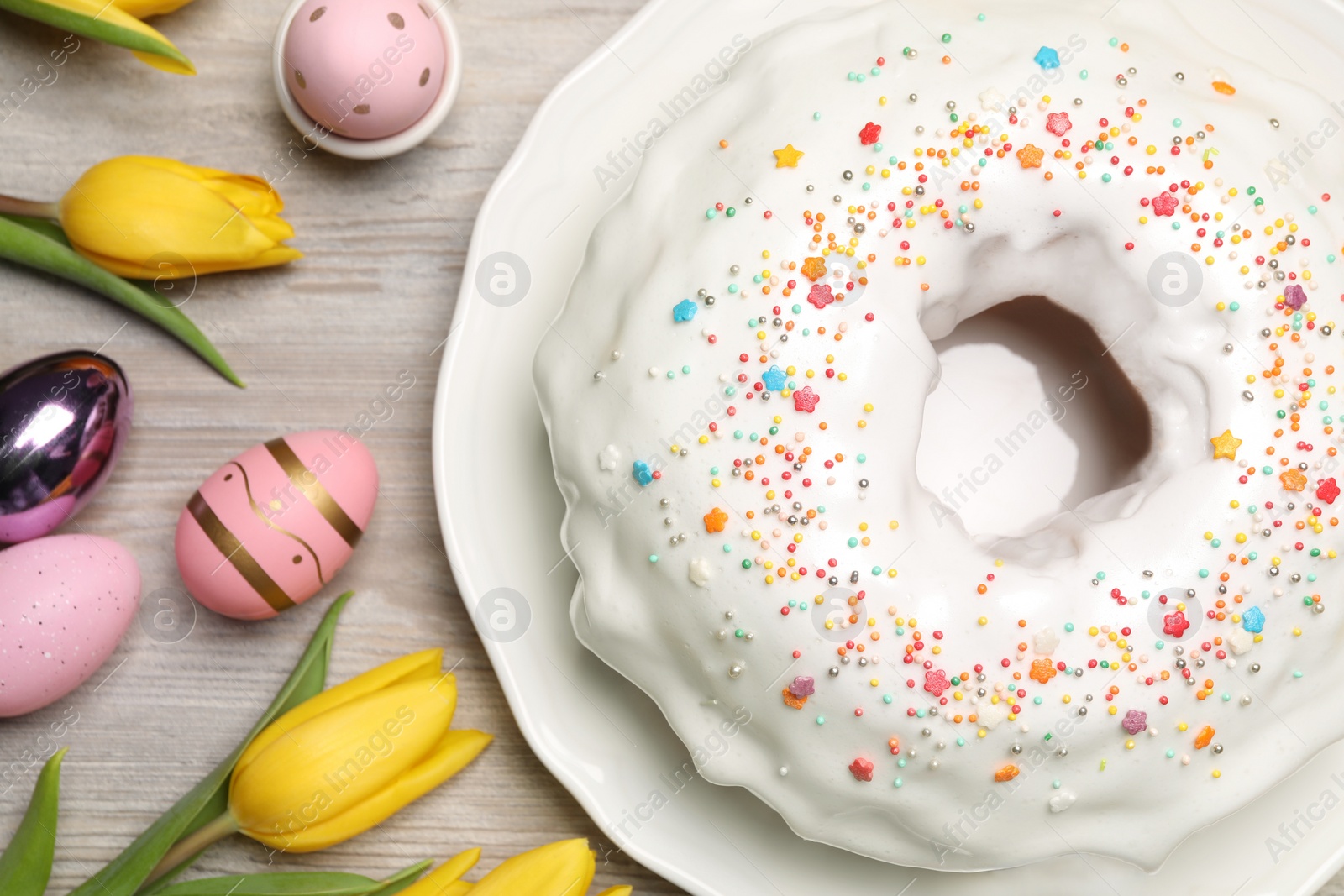 Photo of Easter cake with sprinkles, painted eggs and tulips on white wooden table, flat lay