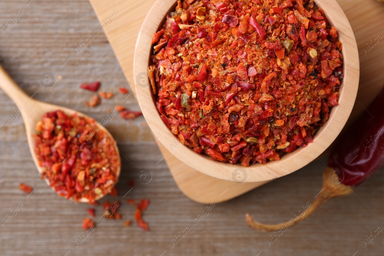 Photo of Aromatic spice. Red chili pepper flakes in bowl, spoon and pod on wooden table, flat lay