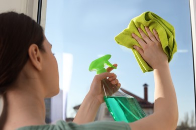 Photo of Young woman cleaning window glass with rag and detergent at home, closeup