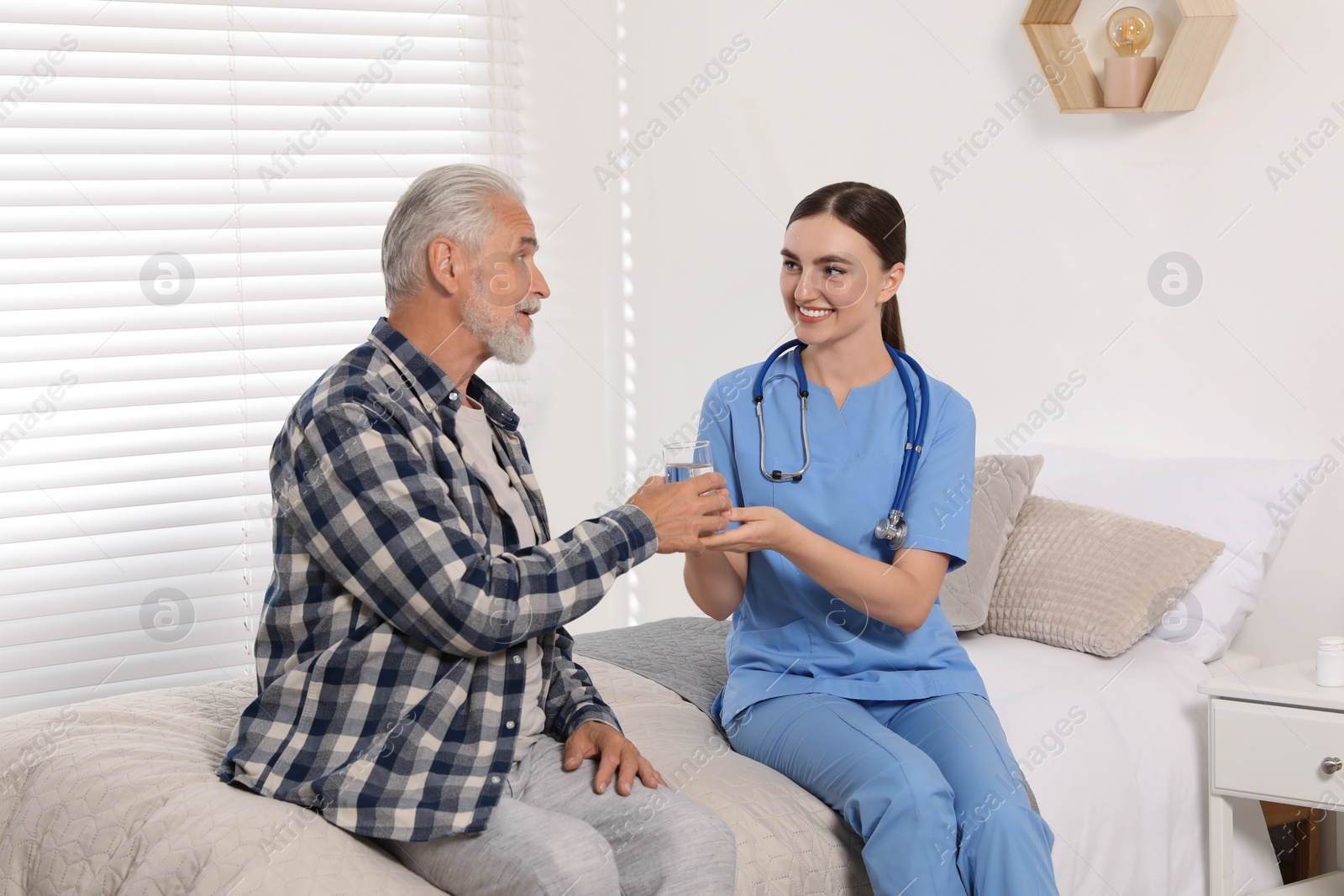 Photo of Young healthcare worker giving glass of water to senior man indoors