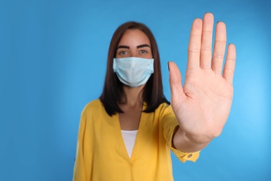 Photo of Young woman in protective mask showing stop gesture on light blue background. Prevent spreading of coronavirus