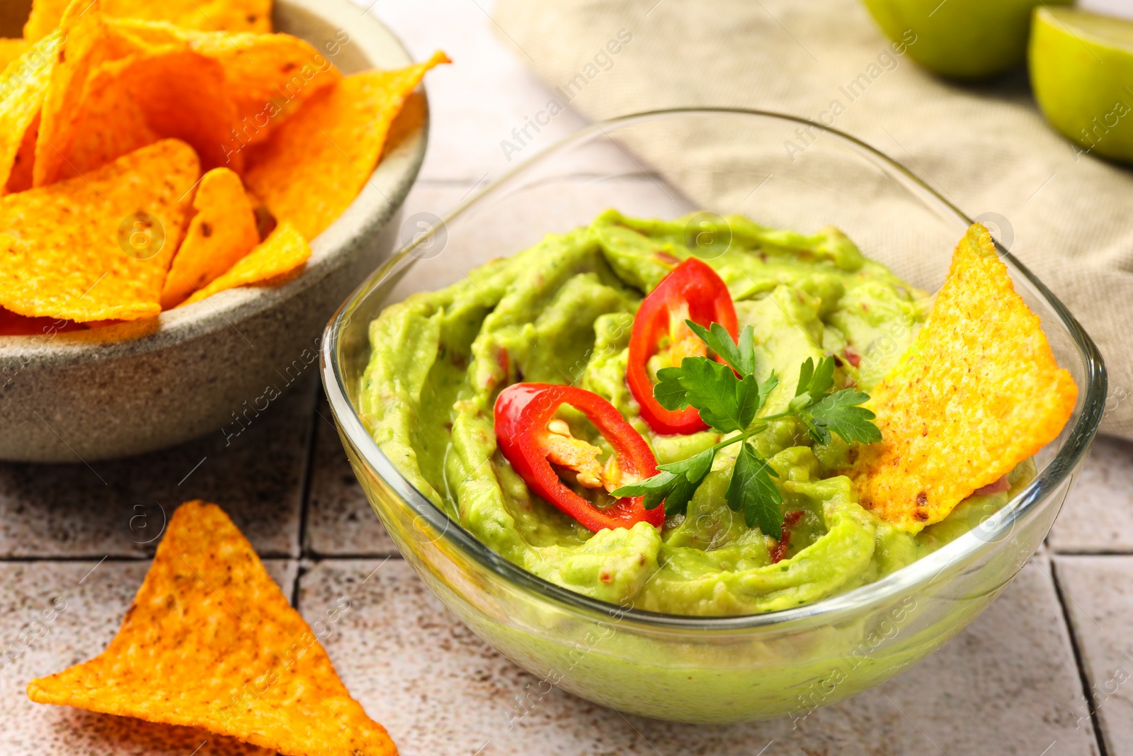 Photo of Bowl of delicious guacamole with chili pepper, nachos chips and lime on white tiled table, closeup