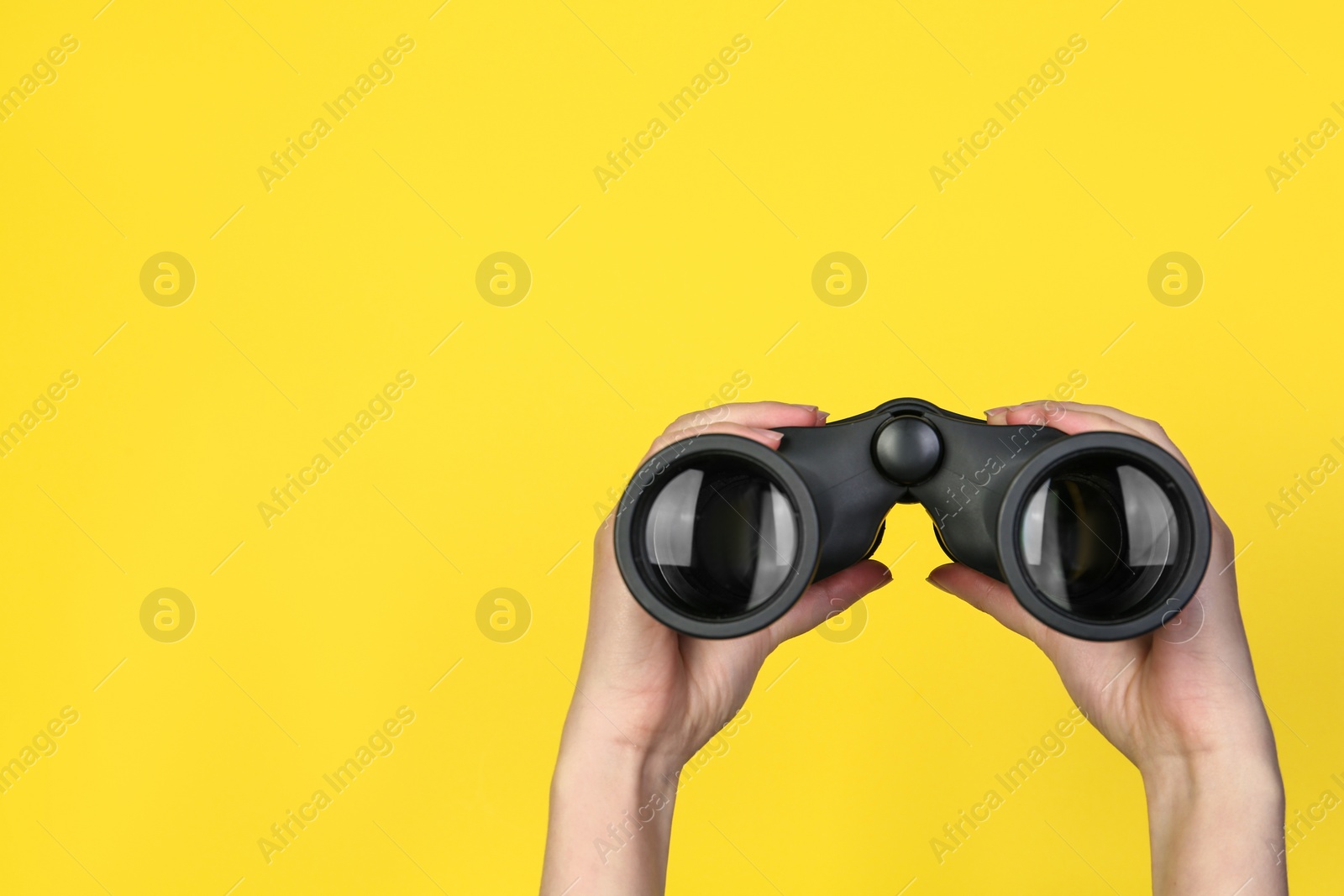 Photo of Woman holding modern binoculars on yellow background, closeup. Space for text