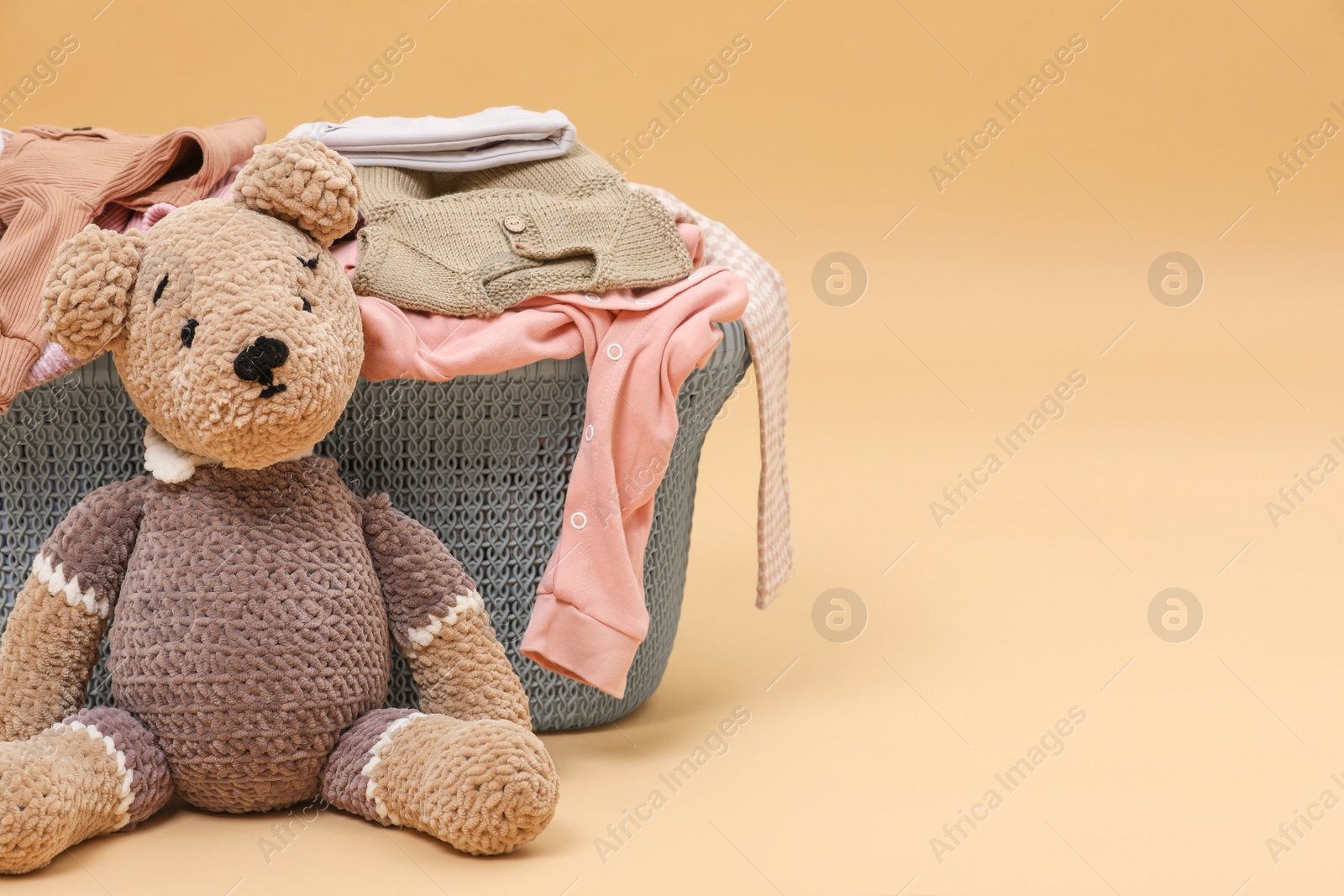 Photo of Laundry basket with baby clothes near soft toy on light brown background. Space for text