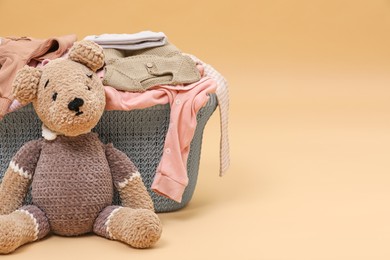 Photo of Laundry basket with baby clothes near soft toy on light brown background. Space for text