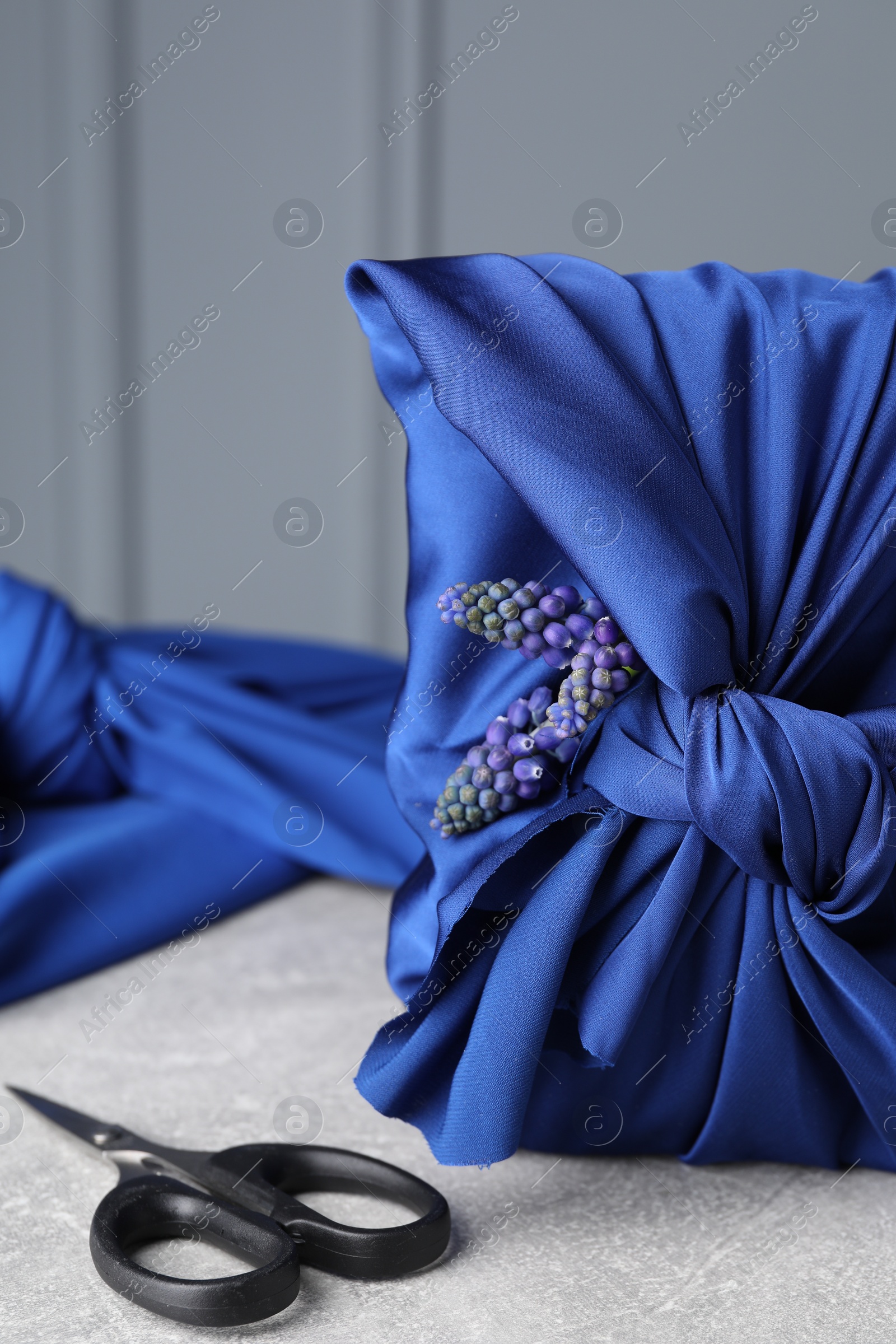 Photo of Furoshiki technique. Gift packed in blue silk fabric, muscari flowers and scissors on light grey table