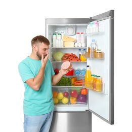 Photo of Young man with expired sausage near open refrigerator on white background