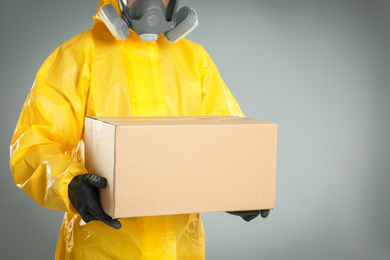 Man wearing chemical protective suit with cardboard box on light grey background, closeup. Prevention of virus spread