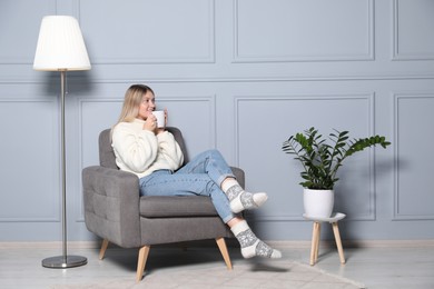 Photo of Young woman with cup of drink relaxing in armchair at home, space for text. Interior design