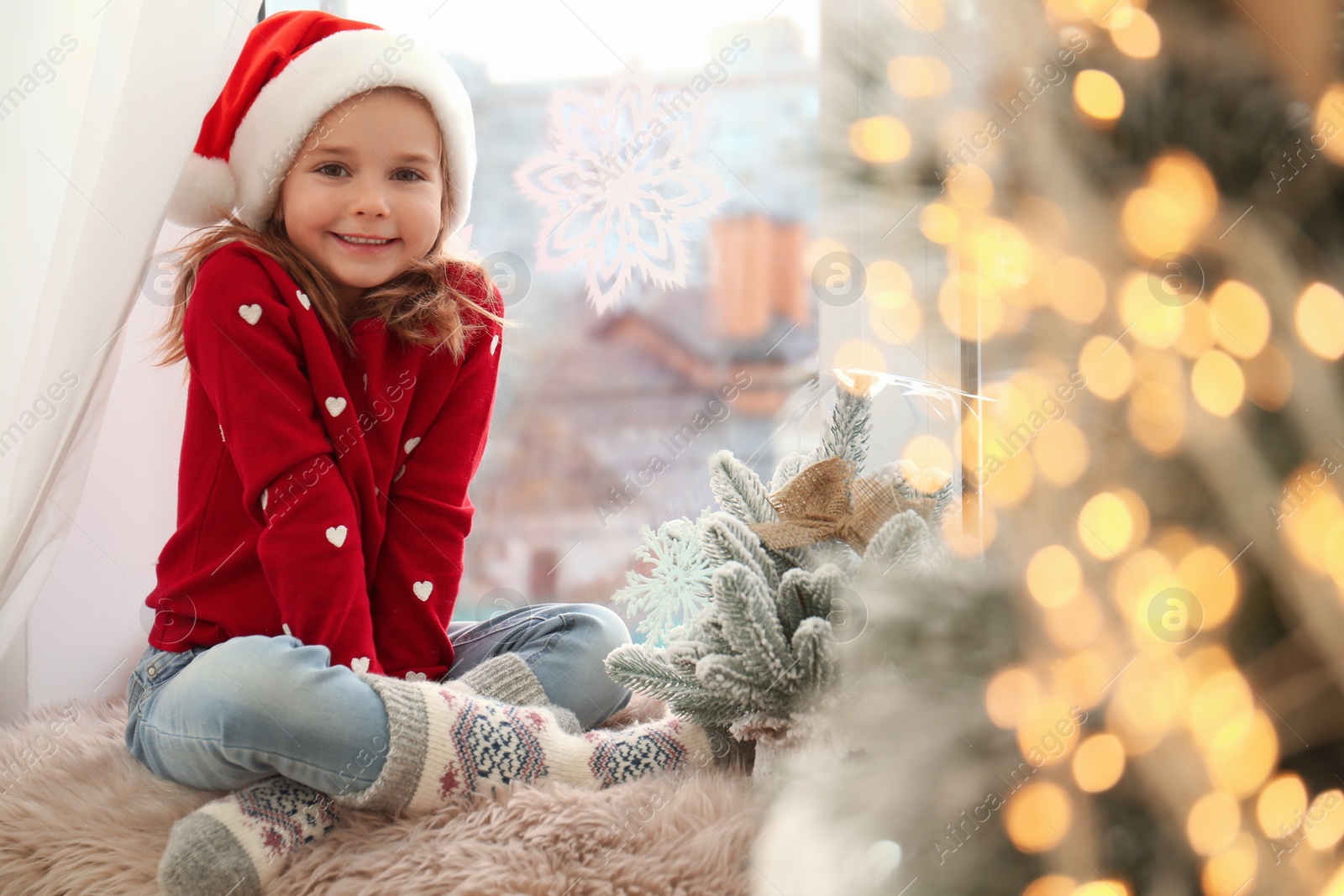 Photo of Little girl in Santa hat near small Christmas tree decorated with snowflakes at home
