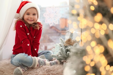 Photo of Little girl in Santa hat near small Christmas tree decorated with snowflakes at home