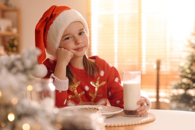 Photo of Cute little girl in Santa hat with delicious Christmas cookie and milk at home, space for text