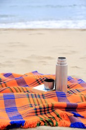 Metallic thermos with hot drink, open book and plaid on sandy beach near sea