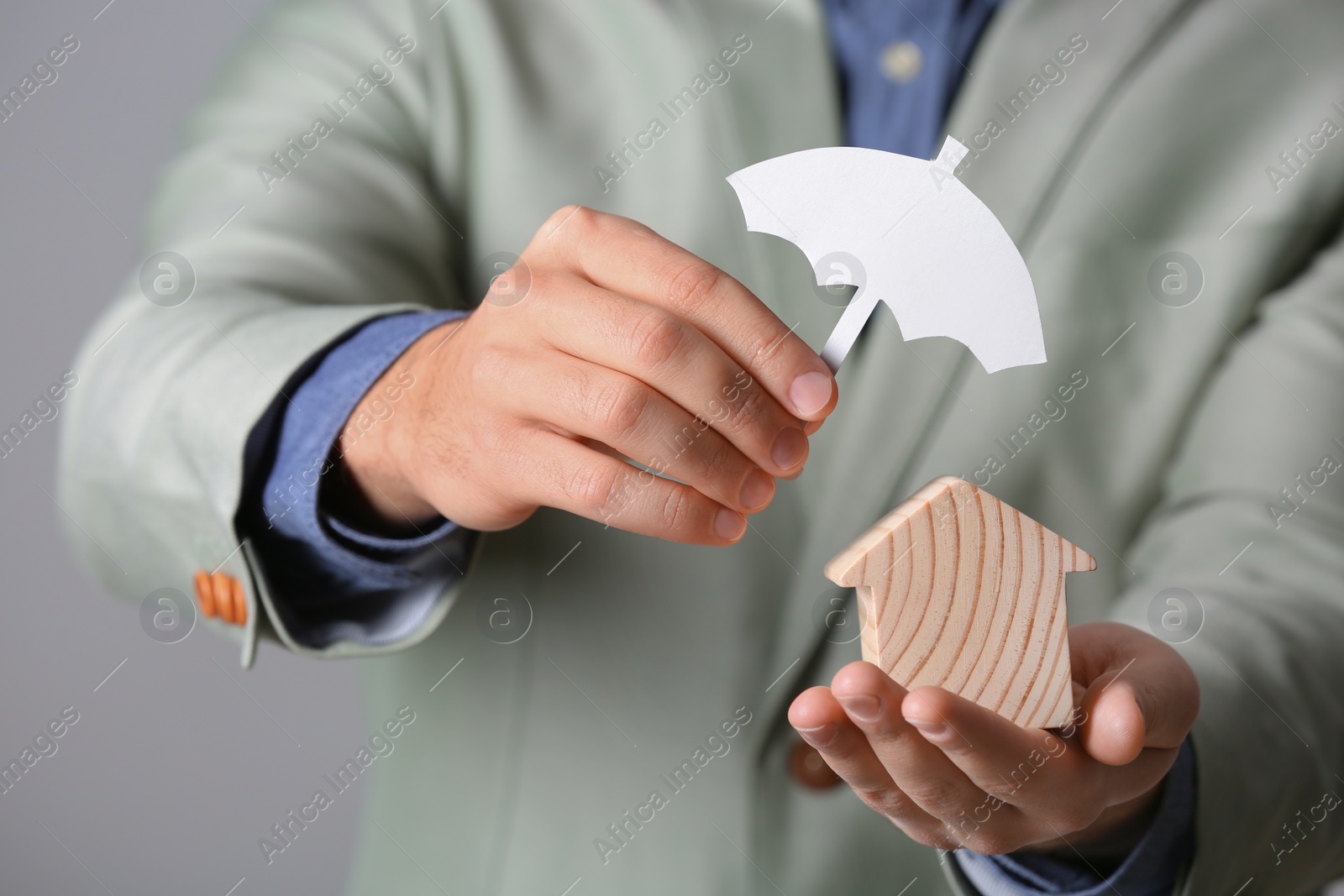 Photo of Male agent covering wooden house with umbrella cutout on grey background, closeup. Home insurance