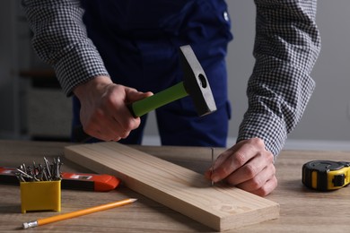 Photo of Professional repairman hammering nail into board at wooden table indoors, closeup