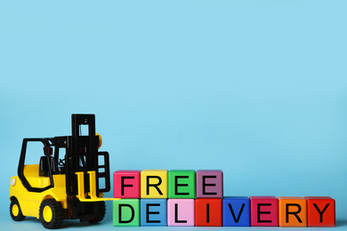 Photo of Toy forklift and cubes with words FREE DELIVERY on light blue background, space for text. Logistics and wholesale concept