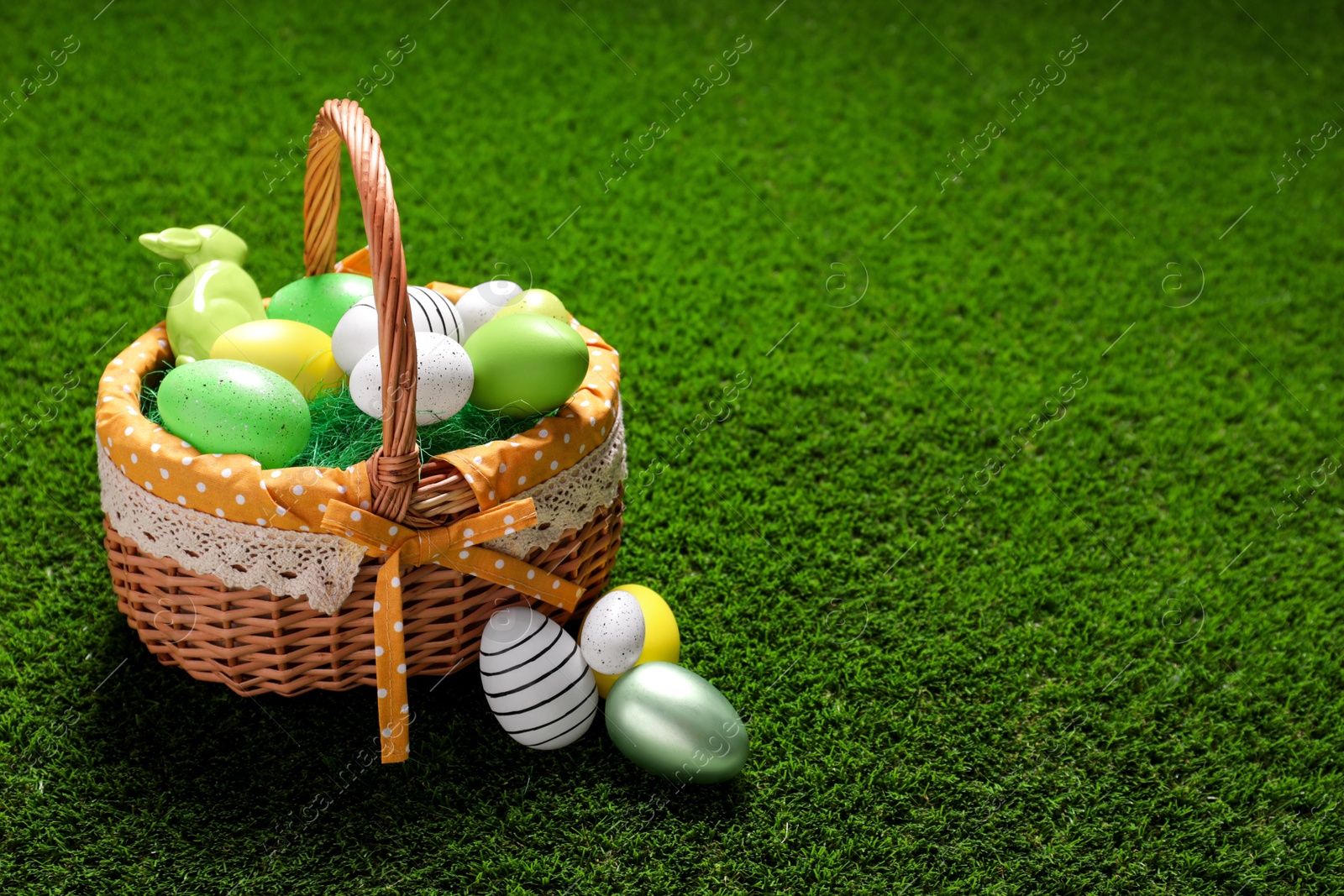 Photo of Easter basket with painted eggs and figure of rabbit on green grass. Space for text