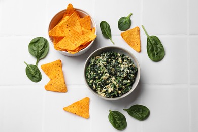 Photo of Tasty spinach dip with egg in bowl and nachos chips on white tiled table, flat lay