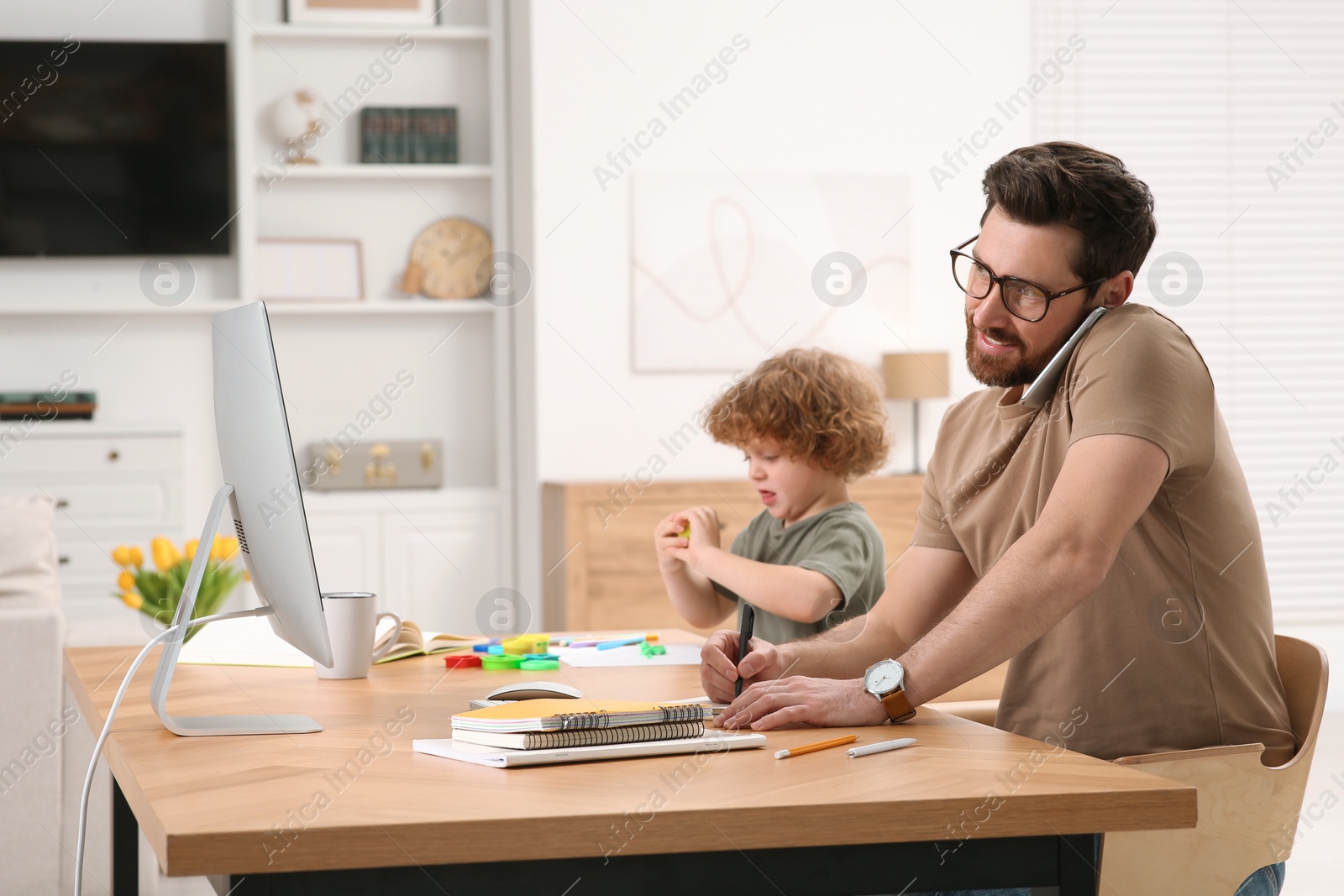 Photo of Man working remotely at home. Busy father talking on smartphone while his son using play dough at desk