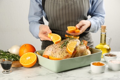 Woman adding orange slices to raw chicken at white marble table, closeup