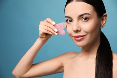 Beautiful young woman doing facial massage with gua sha tool on blue background, closeup