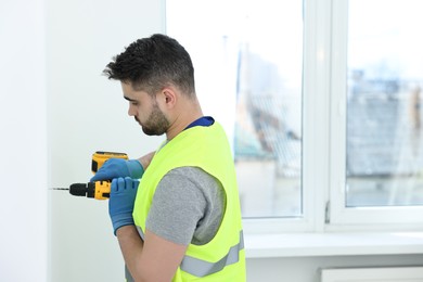 Photo of Young worker in uniform using electric drill indoors. Space for text