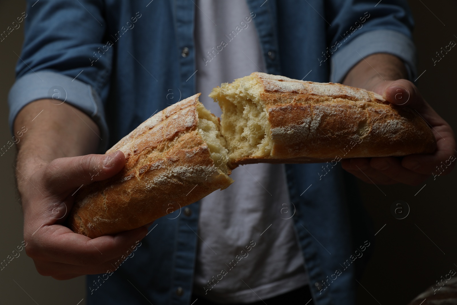 Photo of Man breaking loaf of fresh bread on dark background, closeup