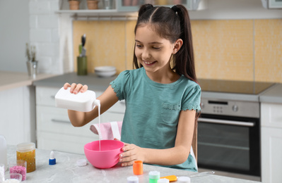 Photo of Cute little girl pouring glue into bowl at table in kitchen. DIY slime toy