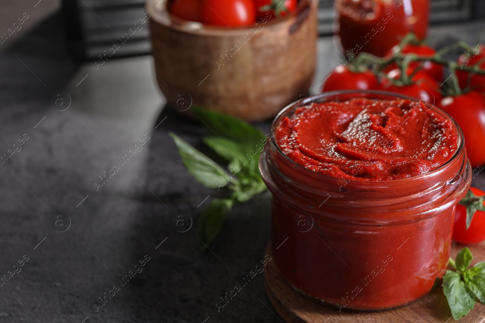 Photo of Jar of tasty tomato paste and ingredients on grey textured table, closeup. Space for text