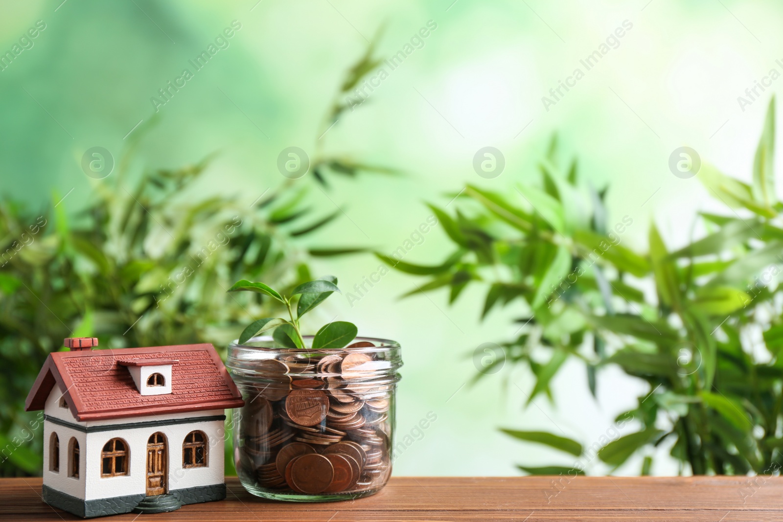 Photo of House model and jar with coins on wooden table against blurred background. Space for text