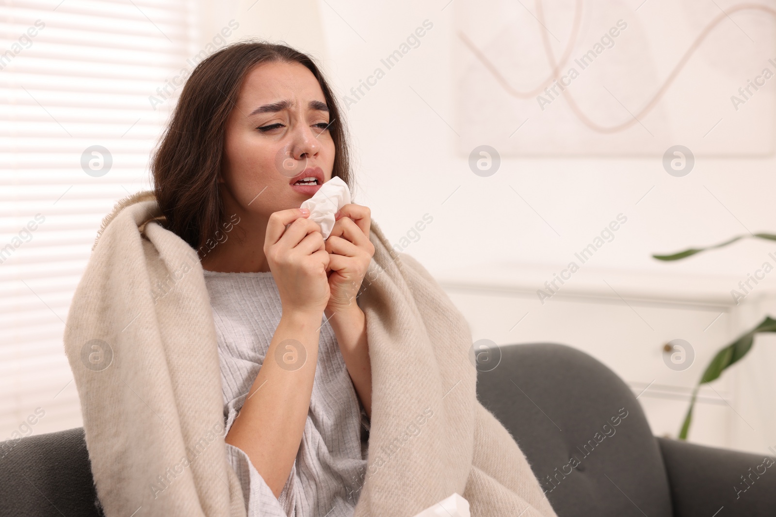 Photo of Sick woman wrapped in blanket with tissue sneezing on sofa at home, space for text. Cold symptoms