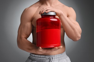 Young man with muscular body holding jar of protein powder on grey background, closeup