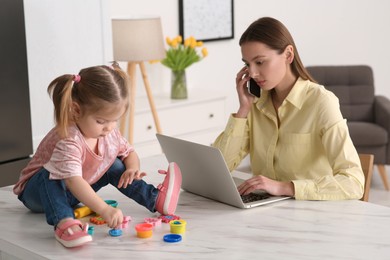 Photo of Woman with laptop working remotely at home. Busy mother talking on phone while her daughter playing on desk