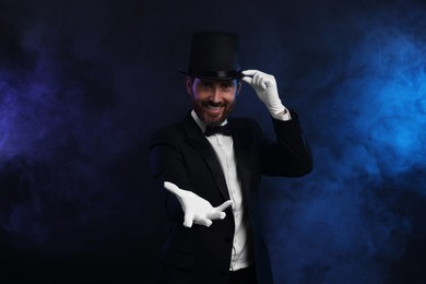 Happy magician wearing top hat in smoke on dark background