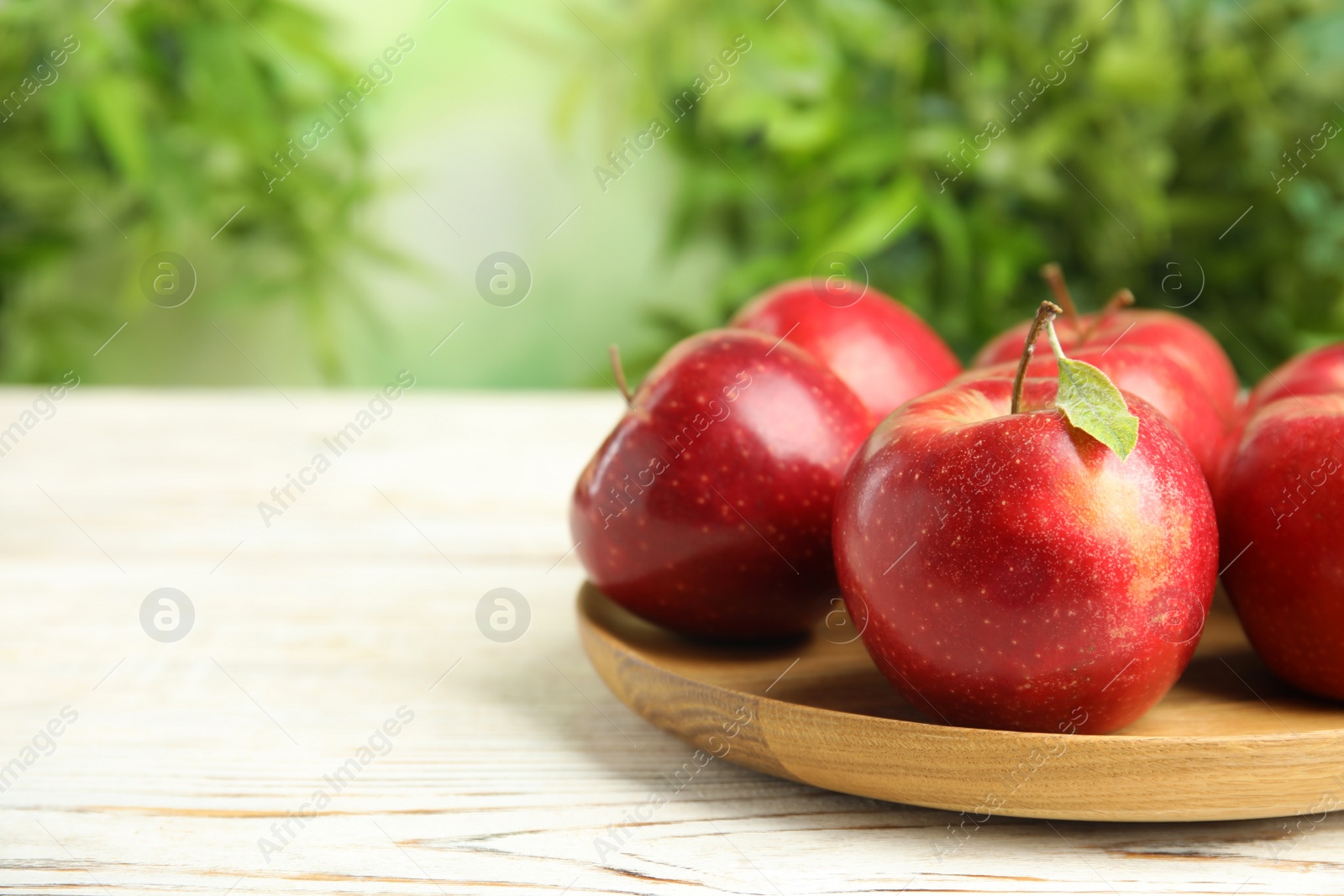 Photo of Wooden plate with ripe juicy red apples on white table against blurred background. Space for text