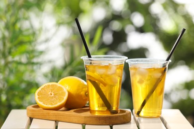 Plastic cups of tasty iced tea with lemon and fresh fruits on white wooden table against blurred green background