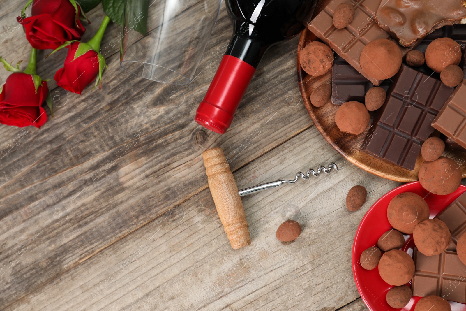 Photo of Bottle of red wine, glass, chocolate sweets, corkscrew and roses on wooden table, flat lay. Space for text