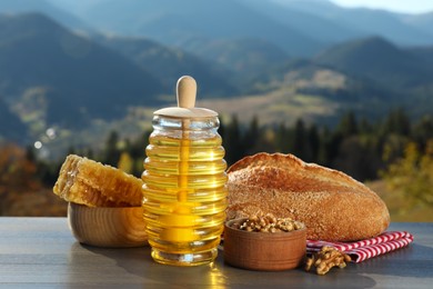 Fresh aromatic honey, combs, bread and nuts on grey wooden table against mountain landscape