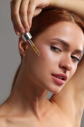 Photo of Beautiful young woman applying cosmetic serum onto her face on grey background, closeup