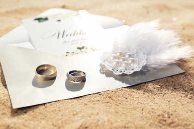 Photo of Envelope with wedding invitation, brooch and gold rings on sandy beach, closeup