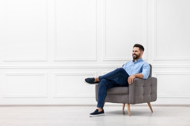 Handsome man sitting in armchair near white wall indoors, space for text