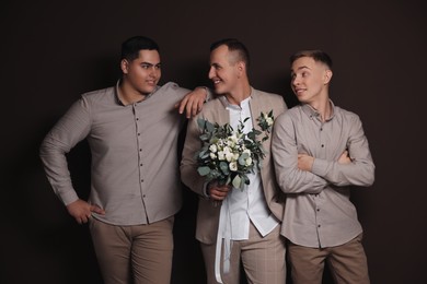 Happy groom with bouquet and his groomsmen on brown background