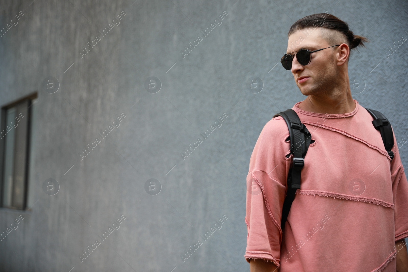 Photo of Handsome young man in stylish sunglasses with backpack near grey wall outdoors, space for text