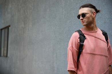 Photo of Handsome young man in stylish sunglasses with backpack near grey wall outdoors, space for text
