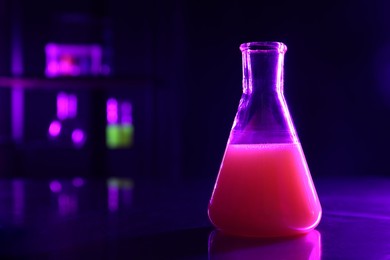 Laboratory flask with luminous liquid on table against dark background, space for text